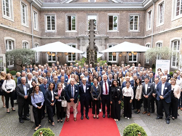 Eine große Gruppe von Menschen steht auf einem roten Teppich in einem Innenhof mit modernen Gebäuden im Hintergrund, umgeben von Pflanzen und Sonnenschirmen, und posiert für ein Gruppenfoto.