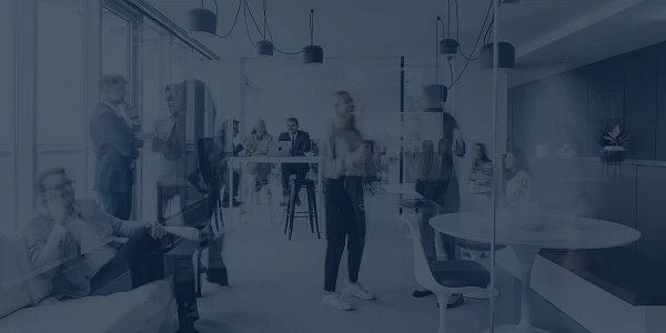 In einem modernen Büroraum unterhalten sich mehrere Personen, während andere an Tischen arbeiten. Der Raum ist hell und stilvoll eingerichtet, mit großen Fenstern und minimalistischen Möbeln. Einige Personen stehen, andere sitzen entspannt auf einer Couch.