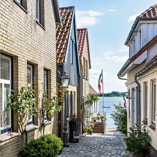Kopfsteinpflastergasse mit Häusern und Blick auf einen See