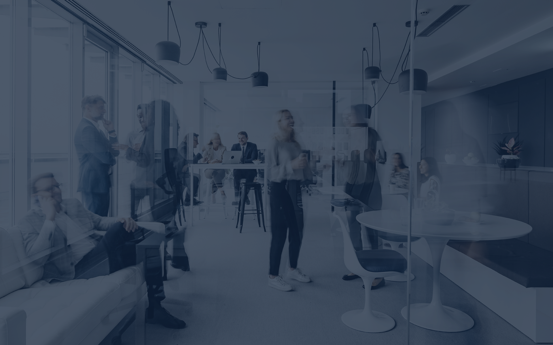 In einem modernen Büroraum unterhalten sich mehrere Personen, während andere an Tischen arbeiten. Der Raum ist hell und stilvoll eingerichtet, mit großen Fenstern und minimalistischen Möbeln. Einige Personen stehen, andere sitzen entspannt auf einer Couch.